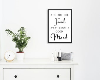 May include: A framed print with the text "YOU ARE ONE Facial AWAY FROM A GOOD Mood" in black script. The print is displayed on a white dresser, alongside a potted plant, a clock, candles, and a small gray basket.