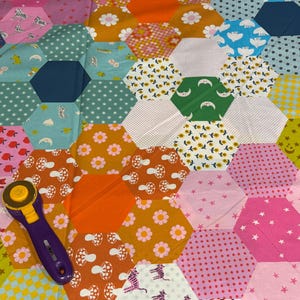 May include: A colorful patchwork quilt with a variety of patterned hexagonal fabric pieces. The quilt features floral, polka dot, and star patterns in shades of pink, orange, green, and blue. A purple rotary cutter with a yellow blade rests on the quilt.