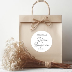 May include: A white round sticker with black text that says "Welcome to the wedding of Olivia + Benjamin 09.28.2024" on a brown paper gift bag with a brown string handle.