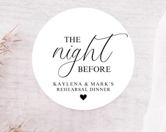 May include: A white circular sticker with the words "THE night BEFORE" in elegant black script. Below, it reads "KAYLENA & MARK'S REHEARSAL DINNER" with a small black heart. The sticker is set against a light wood background.