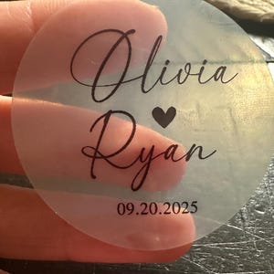May include: A transparent, circular sticker featuring black script. The sticker displays the names "Olivia" and "Ryan", separated by a heart symbol, with the date "09.20.2025" below. The sticker is held by a person.