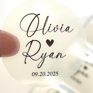 May include: A transparent, circular sticker featuring the names "Olivia" and "Ryan" in cursive script, with a heart symbol between them. The date "09.20.2025" is printed below. Suitable for weddings or similar celebrations.
