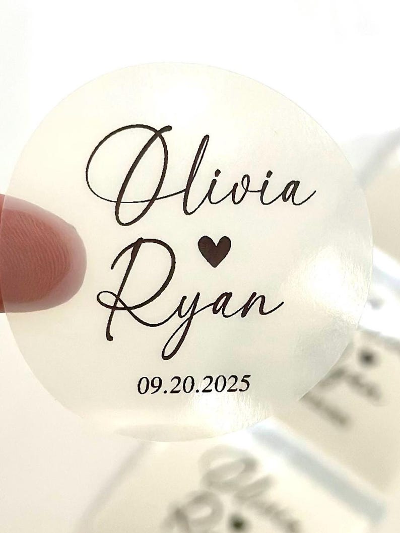May include: A transparent, circular sticker featuring the names "Olivia" and "Ryan" in cursive script, with a heart symbol between them. The date "09.20.2025" is printed below. Suitable for weddings or similar celebrations.