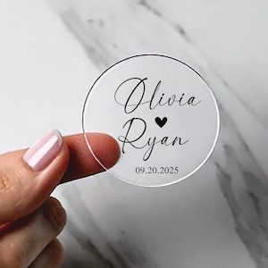May include: A clear, circular wedding favor with black script. The names "Olivia" and "Ryan" are written in elegant cursive, separated by a black heart. The date "09.20.2025" is printed below the names.