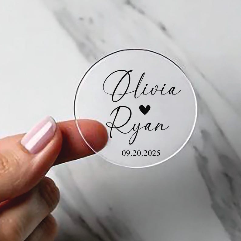 May include: A transparent, circular wedding favour with black script. The names "Olivia" and "Ryan" are written in elegant cursive, separated by a black heart. The date "09.20.2025" is printed below the names.