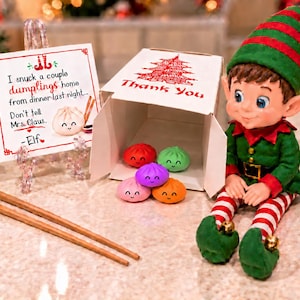 May include: A festive elf doll in a green and red outfit sits beside a white takeout box containing five small, colorful, smiling dumpling figurines. A note reads: I snuck a couple dumplings home from dinner last night... Don't tell Mrs. Claus. - Elf. Two wooden chopsticks lie nearby.
