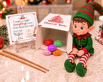 May include: A festive elf doll in a green and red outfit sits beside a white takeout box containing five small, colorful, smiling dumpling figurines. A note reads: I snuck a couple dumplings home from dinner last night... Don't tell Mrs. Claus. - Elf. Two wooden chopsticks lie nearby.