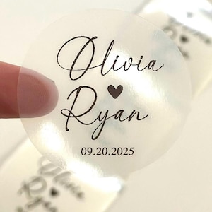 May include: A transparent, circular sticker featuring black script. The text says "Olivia" over "Ryan", with a heart symbol between the names. The date "09.20.2025" is below. The sticker is partially detached from a sheet.