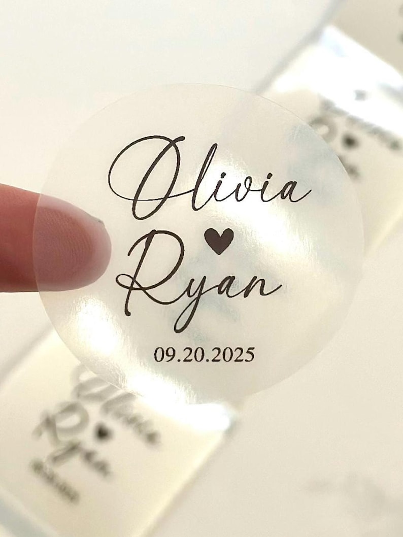 May include: A transparent, circular sticker featuring black script. The text says "Olivia" over "Ryan", with a heart symbol between the names. The date "09.20.2025" is below. The sticker is partially detached from a sheet.