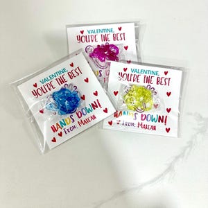 May include: Three Valentine's Day cards with a heart design and the text "Valentine, You're the Best" and "Hands Down!" with a small plastic toy attached to each card.