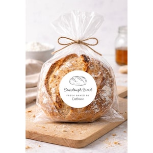 May include: A loaf of sourdough bread in a clear plastic bag tied with twine, sitting on a wooden cutting board. A white sticker on the bag reads "Sourdough Bread, Fresh Baked by Catherine" with a bread illustration.