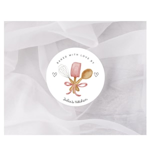 May include: A round sticker with the text "Baked with Love By" and "Julia's Kitchen". The sticker features a whisk, spatula, and wooden spoon tied with a pink ribbon. Two small hearts are on either side of the design.