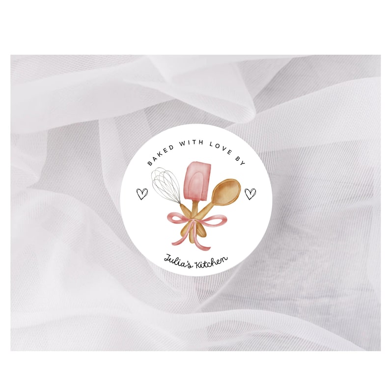 May include: A round sticker with the text "Baked with Love By" and "Julia's Kitchen". The sticker features a whisk, spatula, and wooden spoon tied with a pink ribbon. Two small hearts are on either side of the design.