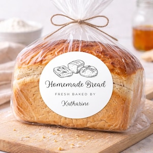 May include: A loaf of homemade bread in a clear plastic bag tied with twine. A white label on the bag reads "Homemade Bread, Fresh Baked by Katharine" with illustrations of bread. The bread sits on a wooden cutting board.