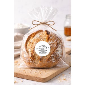 May include: A round loaf of sourdough bread in a clear plastic bag, tied with twine. The bread has a golden-brown crust and is dusted with flour. A white circular label reads "SOURDOUGH MADE WITH LOVE BY STEPHANIE". The bread rests on a wooden cutting board.