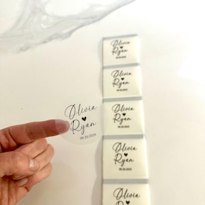 May include: A clear round sticker and white rectangular stickers, each printed with the names "Olivia Ryan" and the date "09.20.2025" in black script. The round sticker is held by a finger. The rectangular stickers are stacked vertically.