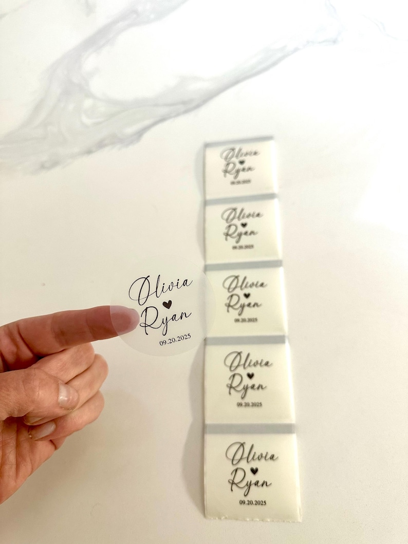 May include: A clear round sticker and white rectangular stickers, each printed with the names "Olivia Ryan" and the date "09.20.2025" in black script. The round sticker is held by a finger. The rectangular stickers are stacked vertically.