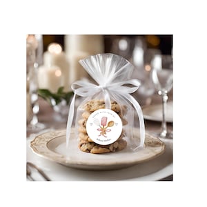 May include: A clear cellophane bag tied with a white ribbon, filled with chocolate chip cookies. A round sticker on the bag reads "Baked with love by Julie's Kitchen" with a whisk, spoon, and heart illustrations. The cookies sit on a white plate.