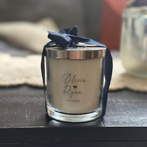 May include: A glass candle with a silver lid and a navy blue ribbon tied in a bow. The candle has the names "Olivia Ryan" and the date "09.20.2025" printed on the front. The candle is on a dark wooden surface.