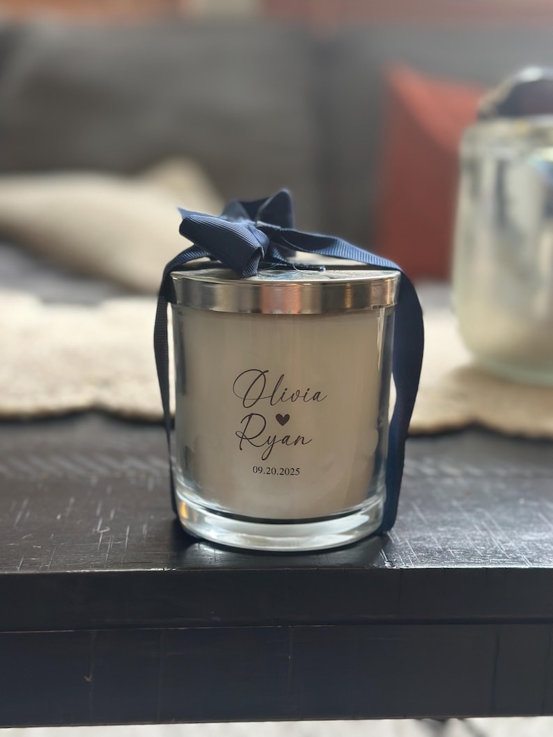 May include: A glass candle with a silver lid and a navy blue ribbon tied in a bow. The candle has the names "Olivia Ryan" and the date "09.20.2025" printed on the front. The candle is on a dark wooden surface.