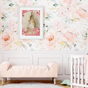 May include: A white crib with a pink and white blanket and a pink and white floral wallpaper with a framed picture of a rabbit holding a teacup.