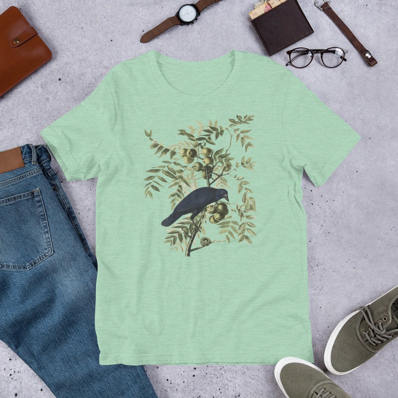 May include: A light green t-shirt with a vintage illustration of a black crow perched on a branch with green leaves and brown berries.