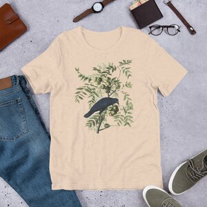 May include: A beige t-shirt with a vintage illustration of a black crow perched on a branch with green leaves and small green fruit.