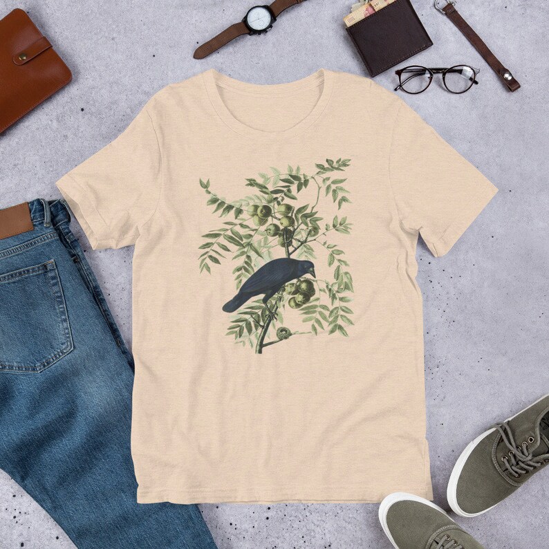 May include: A beige t-shirt with a vintage illustration of a black crow perched on a branch with green leaves and small green fruit.