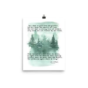 May include: A white art print featuring a watercolor painting of evergreen trees in shades of green and gray. The print includes a quote in black text about gold, wanderers, roots, and kings. The print is held by a silver clip.