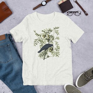 May include: A white t-shirt with a vintage illustration of a black crow perched on a branch with green leaves and fruit.