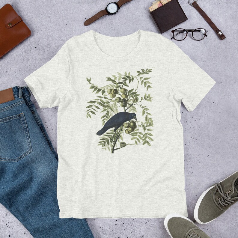 May include: A white t-shirt with a vintage illustration of a black crow perched on a branch with green leaves and fruit.