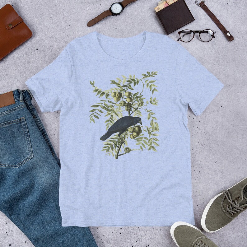 May include: A light blue t-shirt with a vintage illustration of a black crow perched on a branch with green leaves and yellow fruit.