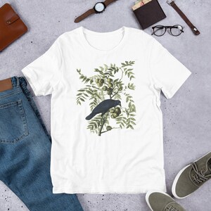 May include: White t-shirt with a black crow perched on a branch with green leaves and small green fruit.