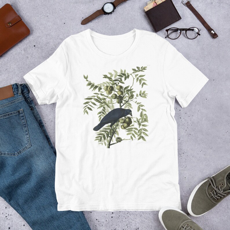 May include: White t-shirt with a black crow perched on a branch with green leaves and small green fruit.