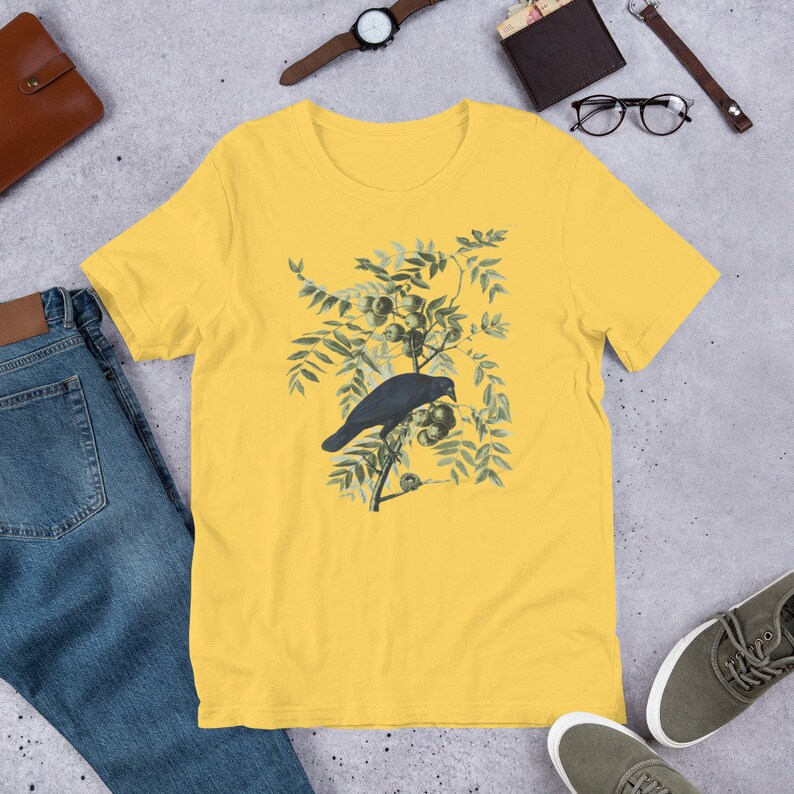 May include: A yellow short-sleeved t-shirt with a vintage illustration of a black crow perched on a branch with green leaves and berries.