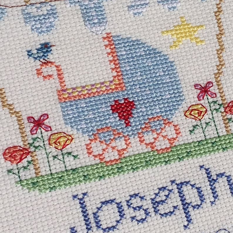 May include: Cross-stitch embroidery of a baby carriage with a blue canopy, a red heart, and a blue bird on top. The carriage is surrounded by flowers and the name "Joseph" is stitched below.