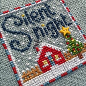 May include: Cross-stitch embroidery of a winter scene with a red house, a green Christmas tree with a yellow star, and the words "Silent Night" in blue thread.