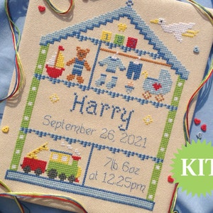 May include: A cross-stitch kit featuring a house with a blue roof and a white bird on top. The house is filled with toys, including a teddy bear, a sailboat, and a toy train. The text "Harry September 26 2021 7lb 6oz at 12:25pm" is stitched below the house. The word "KIT" is written in white on a green circle.