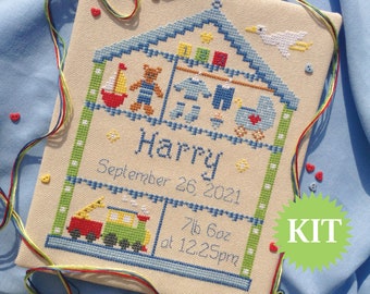 Può includere: Un kit di ricamo a punto croce che rappresenta una casa con un tetto blu e un uccello bianco in cima. La casa è piena di giocattoli, tra cui un orsacchiotto, una barca a vela e un treno giocattolo. Il testo "Harry September 26 2021 7lb 6oz at 12:25pm" è ricamato sotto la casa. La parola "KIT" è scritta in bianco su un cerchio verde.