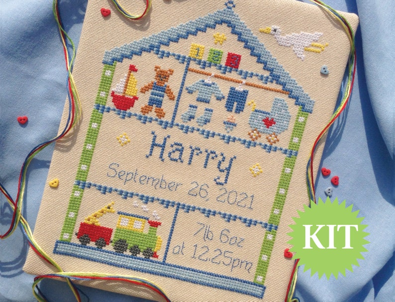 Puede incluir: Un kit de punto de cruz que representa una casa con un techo azul y un p&aacute;jaro blanco en la parte superior. La casa est&aacute; llena de juguetes, incluyendo un oso de peluche, un velero y un tren de juguete. El texto "Harry September 26 2021 7lb 6oz at 12:25pm" est&aacute; bordado debajo de la casa. La palabra "KIT" est&aacute; escrita en blanco sobre un c&iacute;rculo verde.