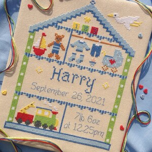 Puede incluir: Una bordado de punto de cruz de una casa con un techo azul y bordes verdes. La casa est&aacute; llena de juguetes, incluyendo un oso de peluche, un velero y un tren. El nombre "Harry" est&aacute; bordado debajo de la casa, junto con la fecha "26 de septiembre de 2021" y el peso y la hora de nacimiento: "7lb 6oz a las 12:25pm".