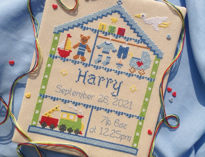Puede incluir: Una bordado de punto de cruz de una casa con un techo azul y bordes verdes. La casa est&aacute; llena de juguetes, incluyendo un oso de peluche, un velero y un tren. El nombre "Harry" est&aacute; bordado debajo de la casa, junto con la fecha "26 de septiembre de 2021" y el peso y la hora de nacimiento: "7lb 6oz a las 12:25pm".