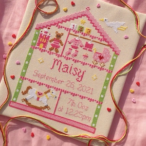 以下が含まれることがあります： ピンクの家のクロスステッチ刺繍。ロッキングホース、テディベア、その他の玩具が描かれています。テキストには「Maisy 2021年9月26日 7lb 0oz 午後12時25分」とあります。