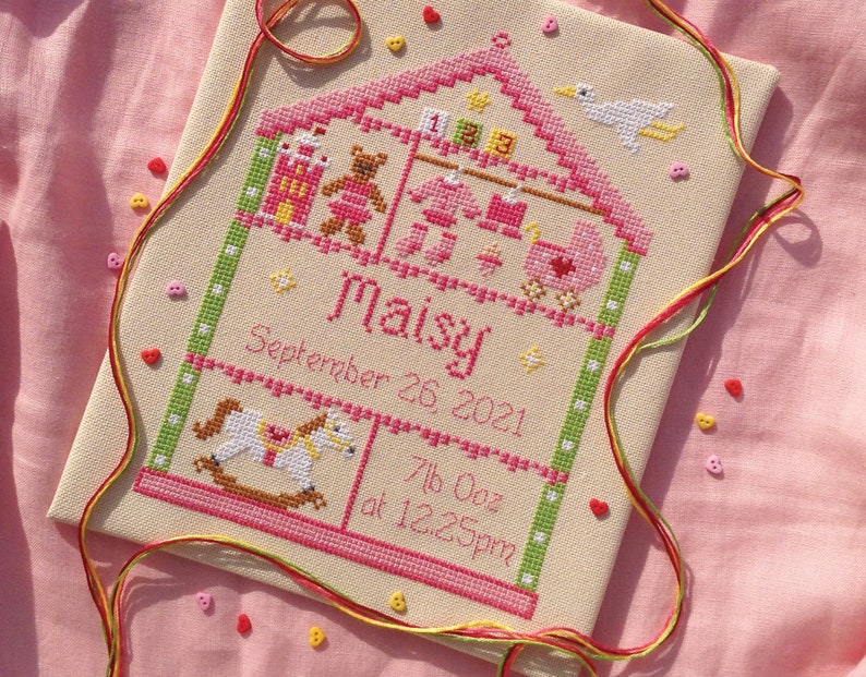 以下が含まれることがあります： ピンクの家のクロスステッチ刺繍。ロッキングホース、テディベア、その他の玩具が描かれています。テキストには「Maisy 2021年9月26日 7lb 0oz 午後12時25分」とあります。
