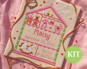 Può includere: Un kit di ricamo a punto croce con una casa rosa e verde con un cavallo a dondolo, un orsacchiotto e un uccello. Il testo sulla casa recita "Maisy Settembre 26, 2021 7lb 0oz alle 12:25 pm".