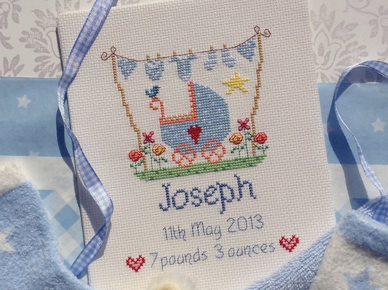 May include: A white cross-stitch embroidery with a blue baby carriage, a yellow star, and red hearts. The text reads "Joseph, 11th May 2013, 7 pounds 3 ounces".