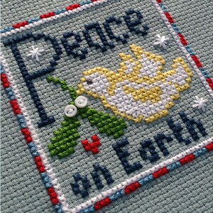 May include: Cross-stitch embroidery with a blue border and a white dove with a green holly sprig and red berries. The text reads "Peace on Earth".