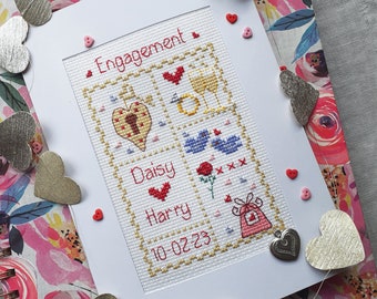 Puede incluir: Una tarjeta blanca con un borde rojo y un diseño de punto de cruz en el interior. El diseño presenta una llave, un anillo, dos pájaros, una rosa y una bolsa de regalo. El texto "Engagement" está en la parte superior de la tarjeta. Los nombres "Daisy" y "Harry" están debajo del diseño, y la fecha "10-02-23" está en la parte inferior.