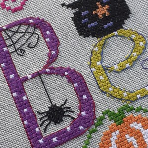 May include: Cross-stitch embroidery of the word "Boo" with a purple "B", a yellow "o", and a black spider hanging from the "B".