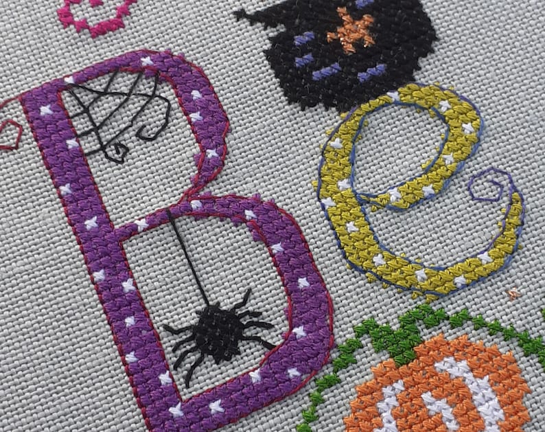 May include: Cross-stitch embroidery of the word "Boo" with a purple "B", a yellow "o", and a black spider hanging from the "B".